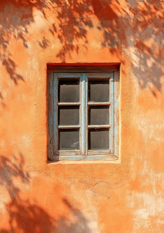 Foto van een oranje terracottamuur met een klein verweerd blauw houten raam en gespikkelde bladschaduwen